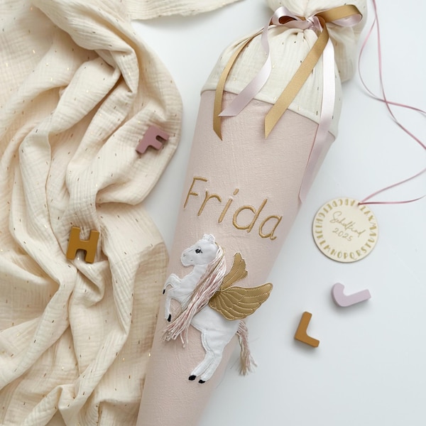 Può includere: Un cono scolastico rosa e oro con un unicorno bianco con ali dorate e il nome "Frida" ricamato su di esso. Il cono è decorato con nastri rosa e dorati e un piccolo cartellino di legno con il testo "Schuleinführung 2023".