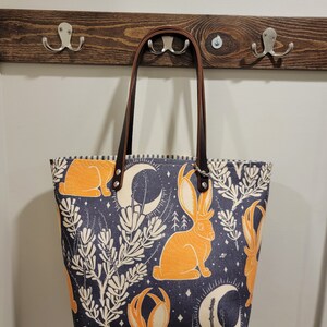 May include: A dark blue tote bag with a brown leather handle and a repeating pattern of orange rabbits, crescent moons, and white floral designs.