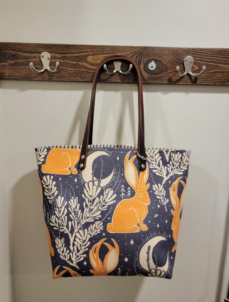 May include: A dark blue tote bag with a brown leather handle and a repeating pattern of orange rabbits, crescent moons, and white floral designs.
