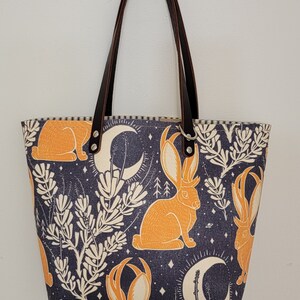 May include: A tote bag with brown leather straps and a dark blue fabric with a repeating pattern of orange rabbits, white crescent moons, and white stylized plants.