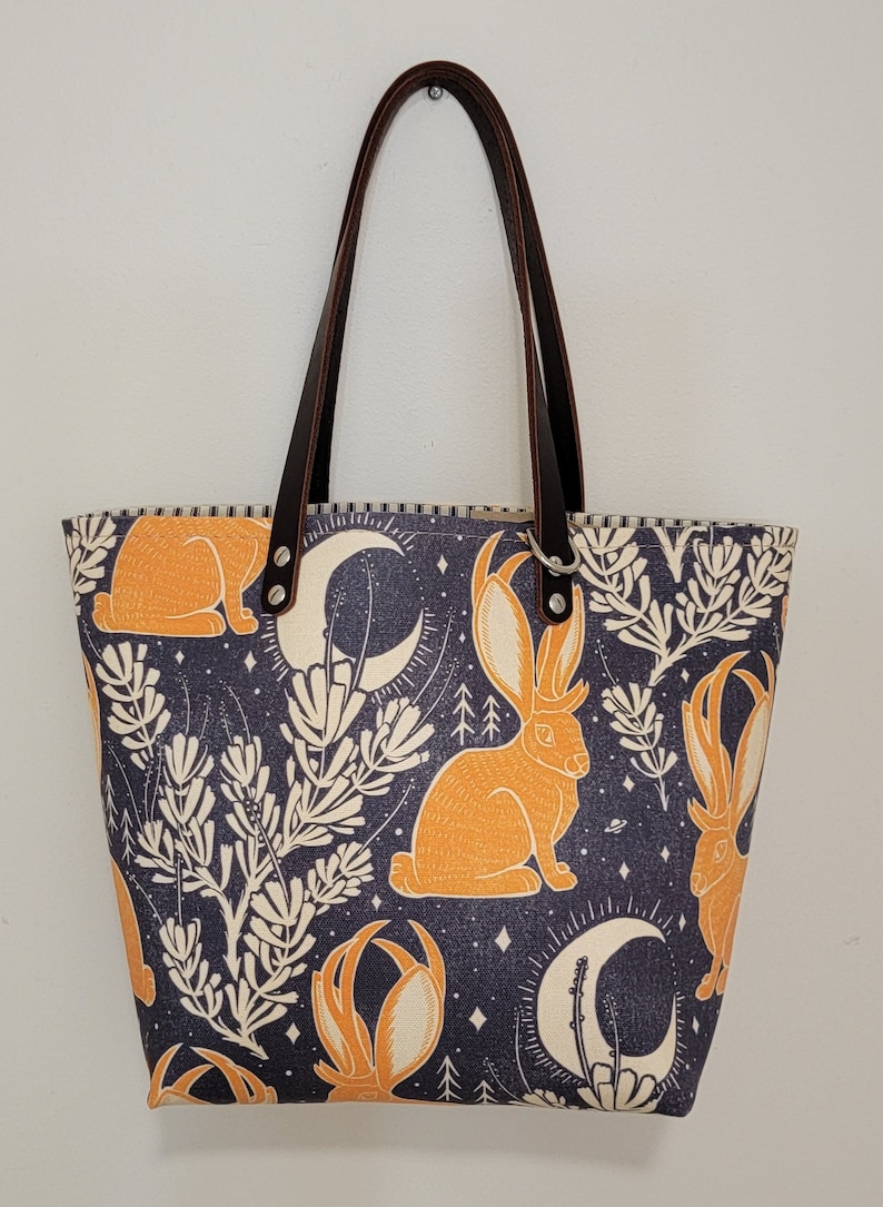 May include: A tote bag with brown leather straps and a dark blue fabric with a repeating pattern of orange rabbits, white crescent moons, and white stylized plants.
