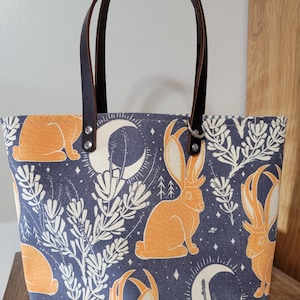 May include: A blue canvas tote bag with brown leather handles. The bag features a repeating pattern of orange rabbits with antlers, white flowers, and crescent moons on a dark blue background.
