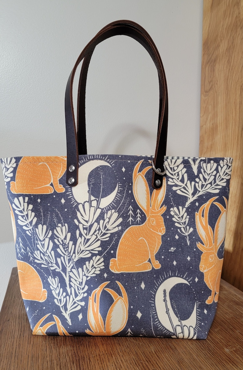 May include: A blue canvas tote bag with brown leather handles. The bag features a repeating pattern of orange rabbits with antlers, white flowers, and crescent moons on a dark blue background.