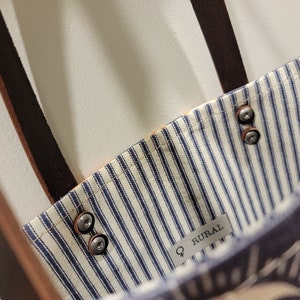 May include: A blue and white striped tote bag with brown leather straps and metal rivets. The bag has a label that says "Q RURAL".