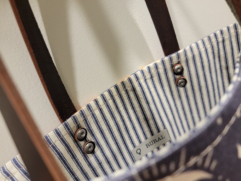 May include: A blue and white striped tote bag with brown leather straps and metal rivets. The bag has a label that says "Q RURAL".