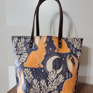 May include: A blue canvas tote bag with brown leather handles. The bag features a repeating pattern of orange rabbits, white crescent moons, and white floral designs on a dark blue background.