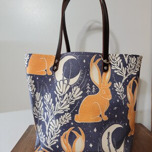 May include: A blue canvas tote bag with brown leather handles. The bag features a repeating pattern of orange rabbits with antlers, white flowers, and crescent moons on a dark blue background.