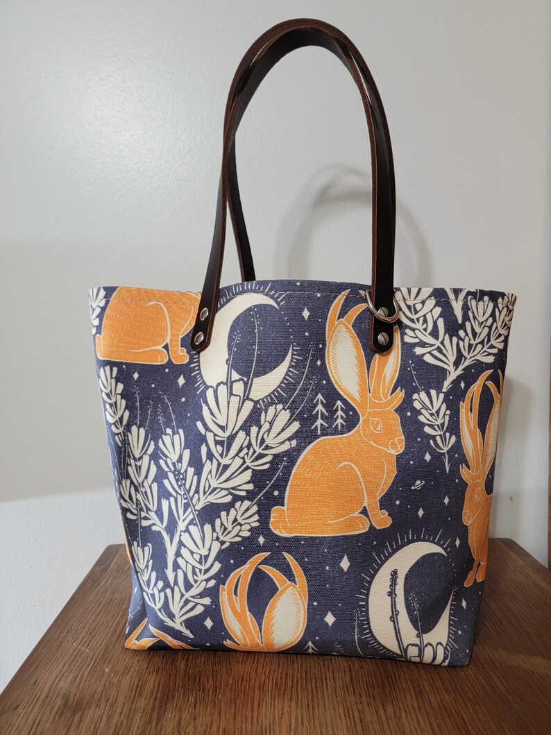 May include: A blue canvas tote bag with brown leather handles. The bag features a repeating pattern of orange rabbits with antlers, white flowers, and crescent moons on a dark blue background.