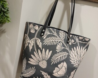 May include: A black tote bag with a floral pattern in a light pink color. The bag has black leather straps and a zipper closure.