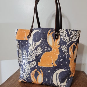 May include: A blue canvas tote bag with brown leather handles. The bag features a repeating pattern of orange rabbits with antlers, white flowers, and crescent moons on a dark blue background.