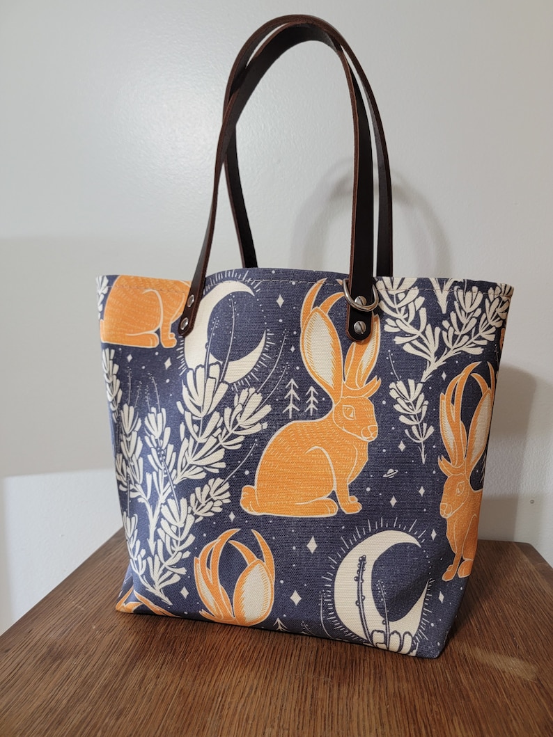 May include: A blue canvas tote bag with brown leather handles. The bag features a repeating pattern of orange rabbits with antlers, white flowers, and crescent moons on a dark blue background.