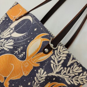 May include: A tote bag with brown leather straps and a navy blue canvas body. The canvas features a repeating pattern of orange rabbits, white flowers, and a crescent moon on a dark blue background.