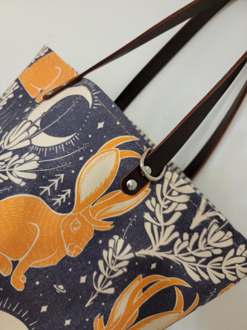 May include: A tote bag with brown leather straps and a navy blue canvas body. The canvas features a repeating pattern of orange rabbits, white flowers, and a crescent moon on a dark blue background.