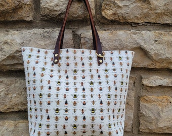 May include: A white tote bag with brown leather handles and a pattern of bees. The bag has a white background with a repeating pattern of bees in various colors and sizes.