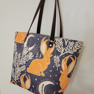 May include: A blue and white tote bag with a pattern of orange rabbits and crescent moons. The bag has brown leather straps.