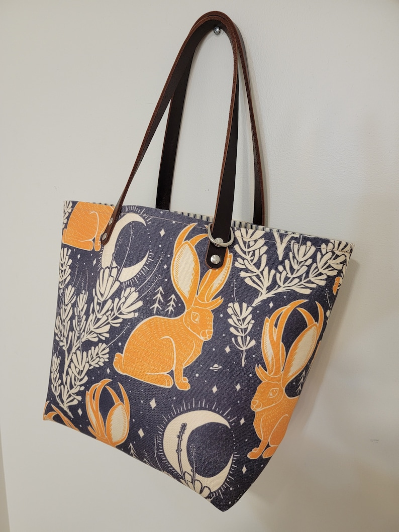 May include: A blue and white tote bag with a pattern of orange rabbits and crescent moons. The bag has brown leather straps.
