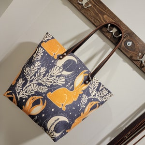 May include: A blue canvas tote bag with a brown leather handle. The bag has a repeating pattern of orange rabbits, white flowers, and crescent moons.