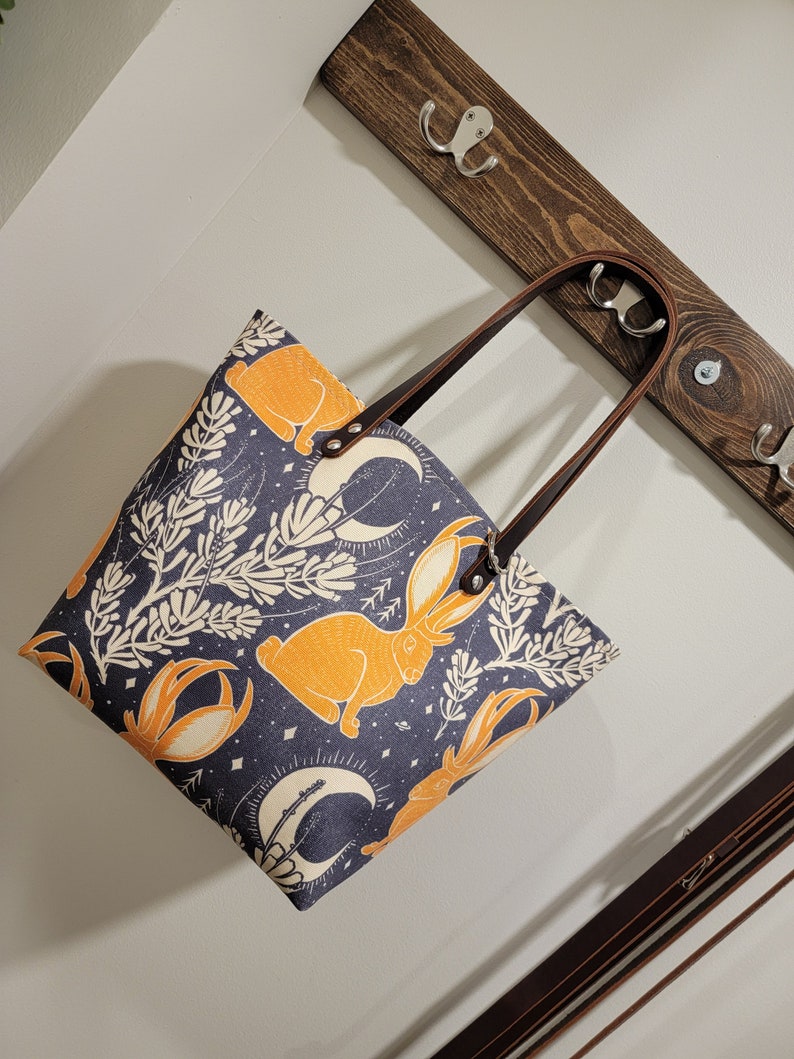 May include: A blue canvas tote bag with a brown leather handle. The bag has a repeating pattern of orange rabbits, white flowers, and crescent moons.