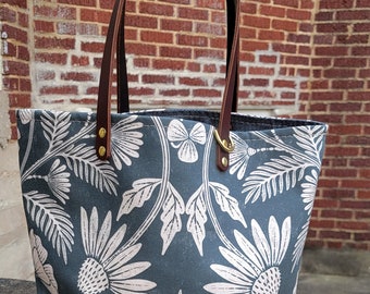 May include: A blue canvas tote bag with white floral print and brown leather handles. The bag has a large, open top and is perfect for carrying everyday essentials.