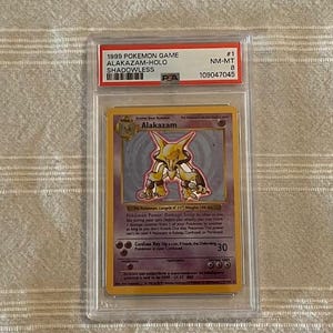 May include: A graded Pokémon card featuring Alakazam, a holo card from the 1999 set. The card is encased in a protective holder with a PSA grade of NM-MT 8. The card has a yellow and purple color scheme.