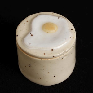 May include: A ceramic jar with a lid featuring a fried egg design. The jar is off-white with brown speckles, and the lid has a white egg white and a yellow yolk. The jar is round and sits on a black surface.