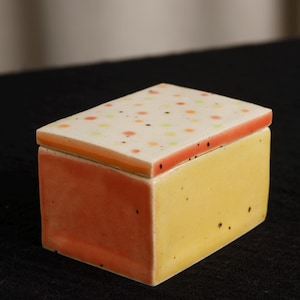 May include: A rectangular ceramic trinket box with a lid. The box is primarily yellow with orange sides. The lid is white with colorful polka dots and an orange border. The box is likely handmade and has a unique, artistic design.