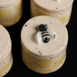 May include: Four small, round ceramic jars with lids. The lids are off-white with a raised bee design. The jars have a yellow and off-white speckled finish. The jars are arranged on a black surface.