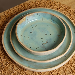 May include: A set of three ceramic plates in varying sizes, nested together. The plates have a light blue glaze with darker speckles and a beige rim. The plates are arranged on a woven, natural-colored placemat.