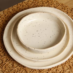 May include: A stack of three off-white ceramic plates and a bowl with a speckled design. The plates and bowl are arranged in a nested fashion, with the bowl at the top. The plates and bowl are on a woven placemat.