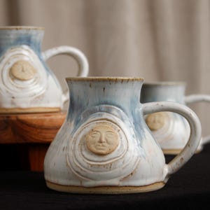 May include: Handmade ceramic mugs with a unique design. Each mug features a sculpted face of the sun, surrounded by concentric circles. The mugs have a blue and white glaze, with a sturdy handle. The mugs are perfect for coffee or tea.
