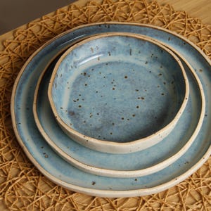May include: A stack of three ceramic plates with a speckled blue glaze and off-white rims. The plates are nested, with a shallow bowl on top, a medium plate in the middle, and a large plate at the bottom. The plates are on a woven mat.
