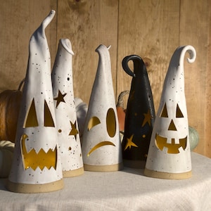 May include: A collection of five tall, conical ceramic ghost lanterns with cut-out facial expressions and star shapes. Four lanterns are white with black speckles, and one is black with yellow stars. Each features a hollow interior with warm light.