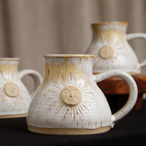 May include: A set of ceramic mugs featuring a white glaze with yellow drip accents at the rim. Each mug has a centered, raised sun face emblem with radiating rays embossed into the clay body. The mugs have a wide base and a curved handle.