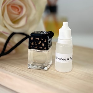 May include: A black and clear glass car air freshener with a black metal cage and a black string. The air freshener is sitting on a wooden tray with a small bottle of clear liquid labelled "Lychee & Peach".