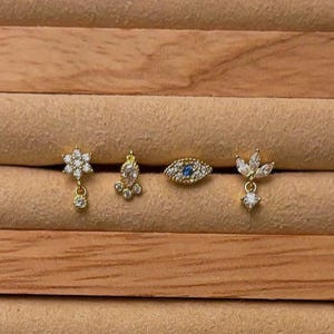 Könnte beinhalten: Vier goldfarbene Ohrringe mit klaren und blauen Kristallen. Die Ohrringe haben ein Blumen-, ein Tropfen-, ein böses Auge- und ein Blattdesign.