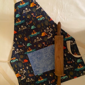 May include: A blue and white patterned apron with a large pocket and a brown rolling pin. The apron features a camping theme with illustrations of animals, trees, and tents.