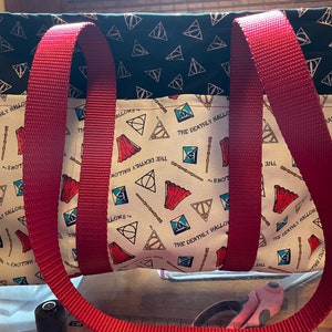 May include: A red and black tote bag with a Harry Potter theme. The bag has a black fabric with gold Deathly Hallows symbols and a white fabric with the words "The Deathly Hallows" and the Deathly Hallows symbols in red, blue, and gold.