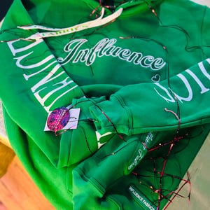 May include: Green hoodie with white drawstring and the word "Influence" in cursive across the chest. The sleeves have the word "Influence" printed in white. The hoodie has a tag that says "Influence Fabrics".