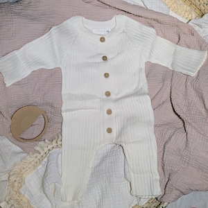 May include: Cream-colored baby romper with long sleeves and wooden buttons down the front. The ribbed knit fabric is soft and comfortable. A wooden sun-shaped teether is also visible.