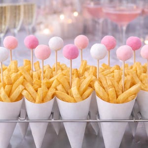May include: Golden french fries in white paper cones, topped with pink and white cake pops on wooden sticks. The cones are arranged on a metal tray, with champagne and pink cocktails in the background.