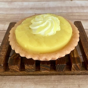 May include: A decorative soap shaped like a lemon tart, featuring a yellow filling and a swirl of white cream on top. The tart sits on a dark brown wooden soap dish with a slatted design.