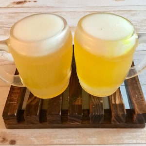 May include: Two clear glass beer mugs filled with yellow liquid and topped with white foam, resting on a dark brown wooden coaster. The mugs have handles and are positioned side-by-side.