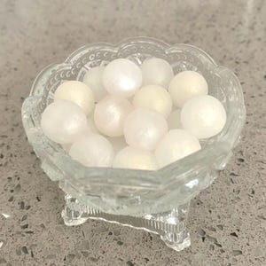 May include: A clear, scalloped-edge glass bowl filled with white, pearl-like spheres. The bowl has a pedestal base and is sitting on a speckled gray surface. The spheres have a slight shimmer.