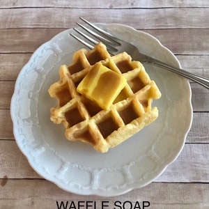 May include: A square waffle-shaped soap with a pat of yellow soap on top, presented on a white plate with a decorative edge. A silver fork rests on the plate. The text "WAFFLE SOAP" is at the bottom.