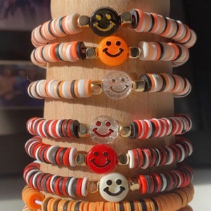 May include: A stack of colorful beaded bracelets with smiley face charms. The bracelets are made with orange, red, black, and white beads.