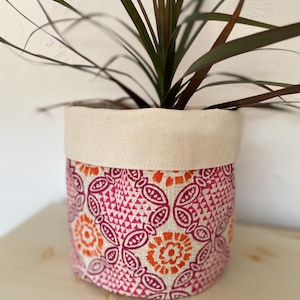 May include: A pink and orange patterned fabric planter with a white canvas top. The planter is filled with a green plant with long, dark green leaves.