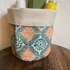 May include: A white fabric storage bin with a teal and orange geometric pattern. The bin is filled with various items, including a white bottle, a green box labelled "Lemon Toner", and a yellow spray bottle.