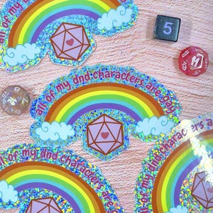 Könnte beinhalten: Regenbogenfarbene Aufkleber mit dem Text "all of my dnd characters are gay" und einem geometrischen Herz-Symbol. Würfel mit Zahlen sind ebenfalls im Bild zu sehen.