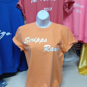 May include: A light orange t-shirt with the text "Scripps Ranch" in white cursive lettering.