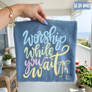 May include: A blue jean colored sweatshirt with the words "Worship While You Wait" in pastel colors. The sweatshirt is held up by a hand with a gold bracelet. The brand name "Sweet Cheeks Apparel & More" is printed on the bottom right.